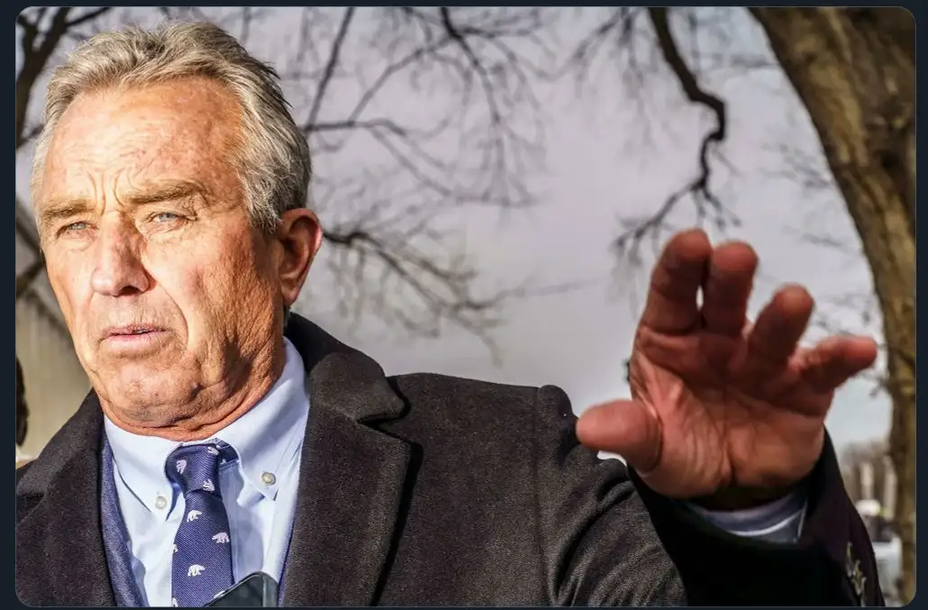 a close-up photo of an older man with gray hair, wearing a dark coat, light blue shirt, and a purple tie with a bear…