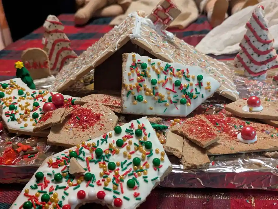 A gingerbread house decorated with white icing, red and green sprinkles, and candy pieces is shown collapsed and shattered…