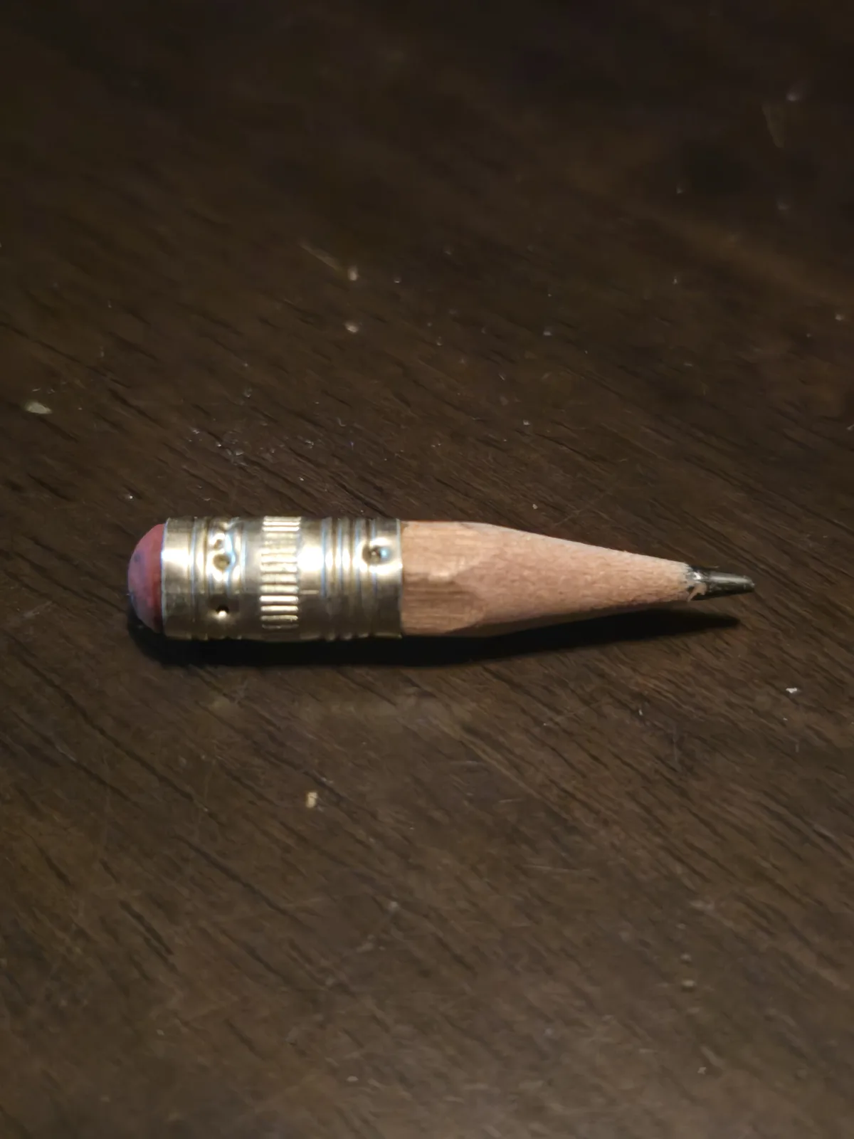 A pencil with its eraser and metal ferrule intact lies on a dark wooden surface, but its wooden body has been shaved down to…