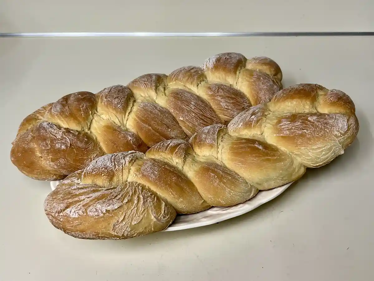 A white plate holds three large, golden-brown braided loaves of bread, dusted with flour, arranged on a plain white…