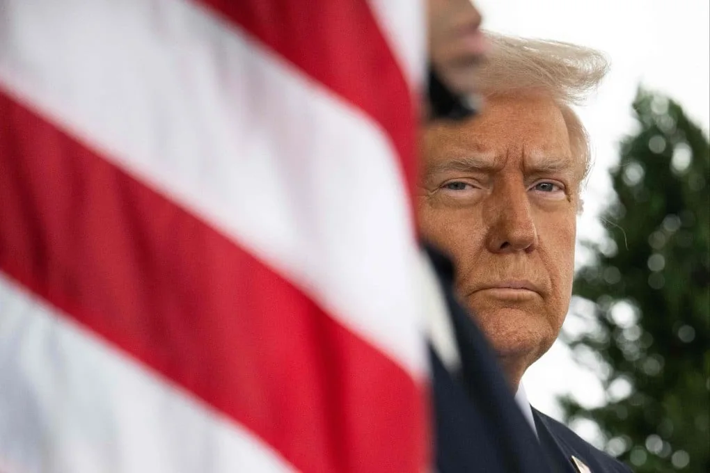 a close-up photo shows former president donald trump with a stern expression, partially obscured by a blurred american flag…