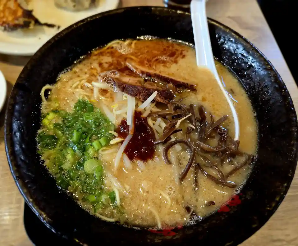 A close-up photo shows a steaming bowl of ramen in a dark ceramic bowl, featuring noodles, broth, sliced pork, bean sprouts…