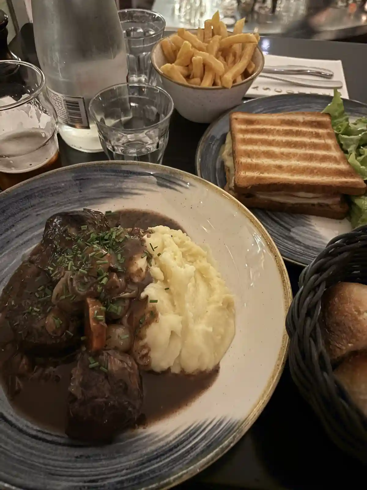 A close-up photo of a restaurant table with a plate of beef bourguignon with mashed potatoes, a grilled sandwich, fries, and…
