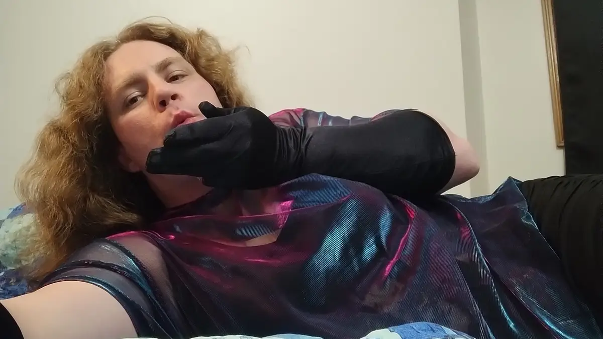 A person with curly hair lies on a couch wearing a shiny, iridescent top and black gloves, holding their hand to their mouth…