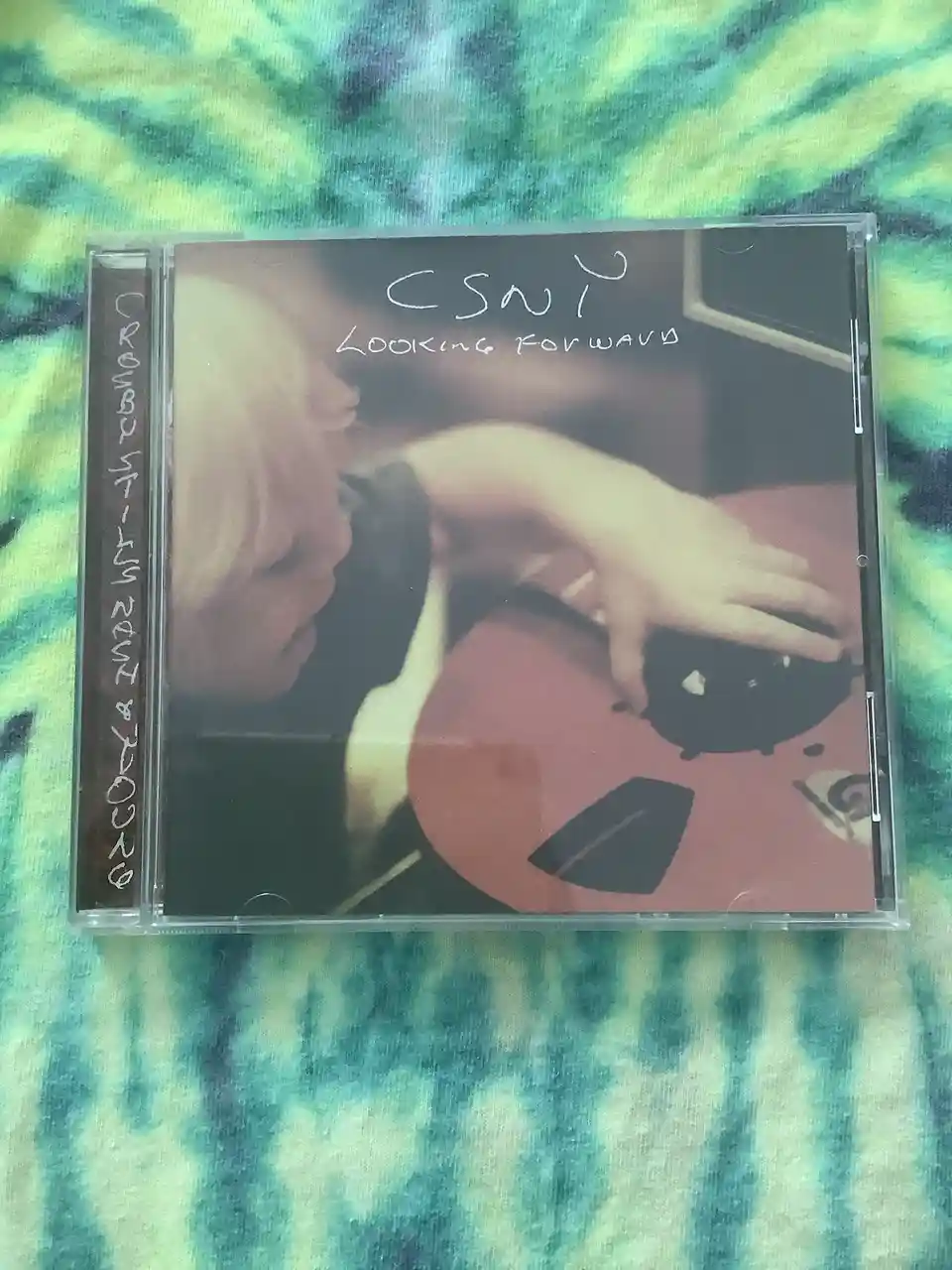 A CD case for the CSNY album 'Looking Forward' is shown on a tie-dye fabric. The cover art features a person with blonde…