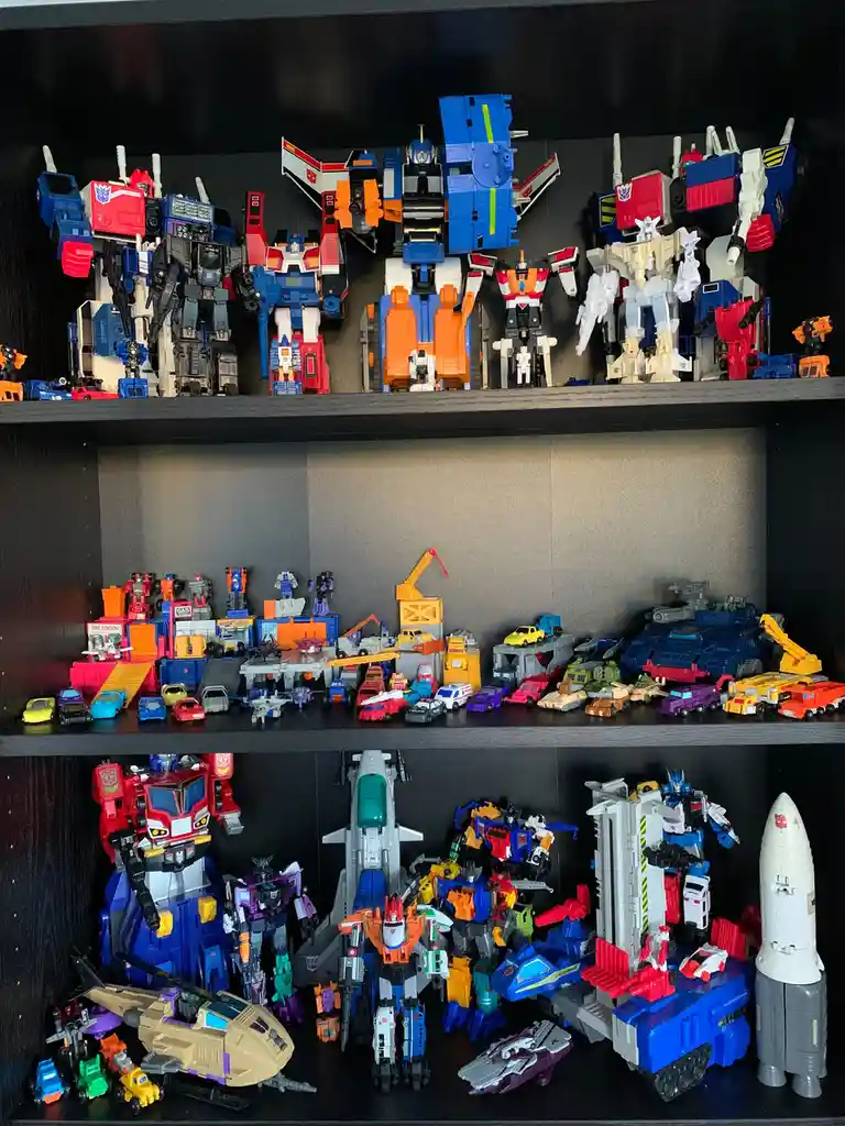 Three shelves display a large collection of Transformers action figures and vehicles, including giant robot figures, smaller…