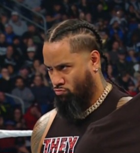 A close-up of wrestler Roman Reigns in a wrestling ring, looking intensely at the camera with a serious expression. He has…