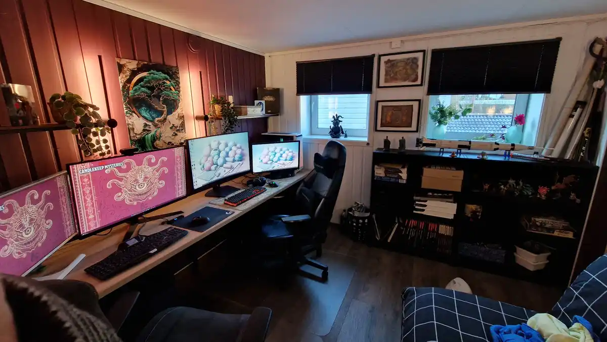 A cozy, well-lit home office features a long desk with four computer monitors, one displaying a pink 'Candlekeep Mysteries'…