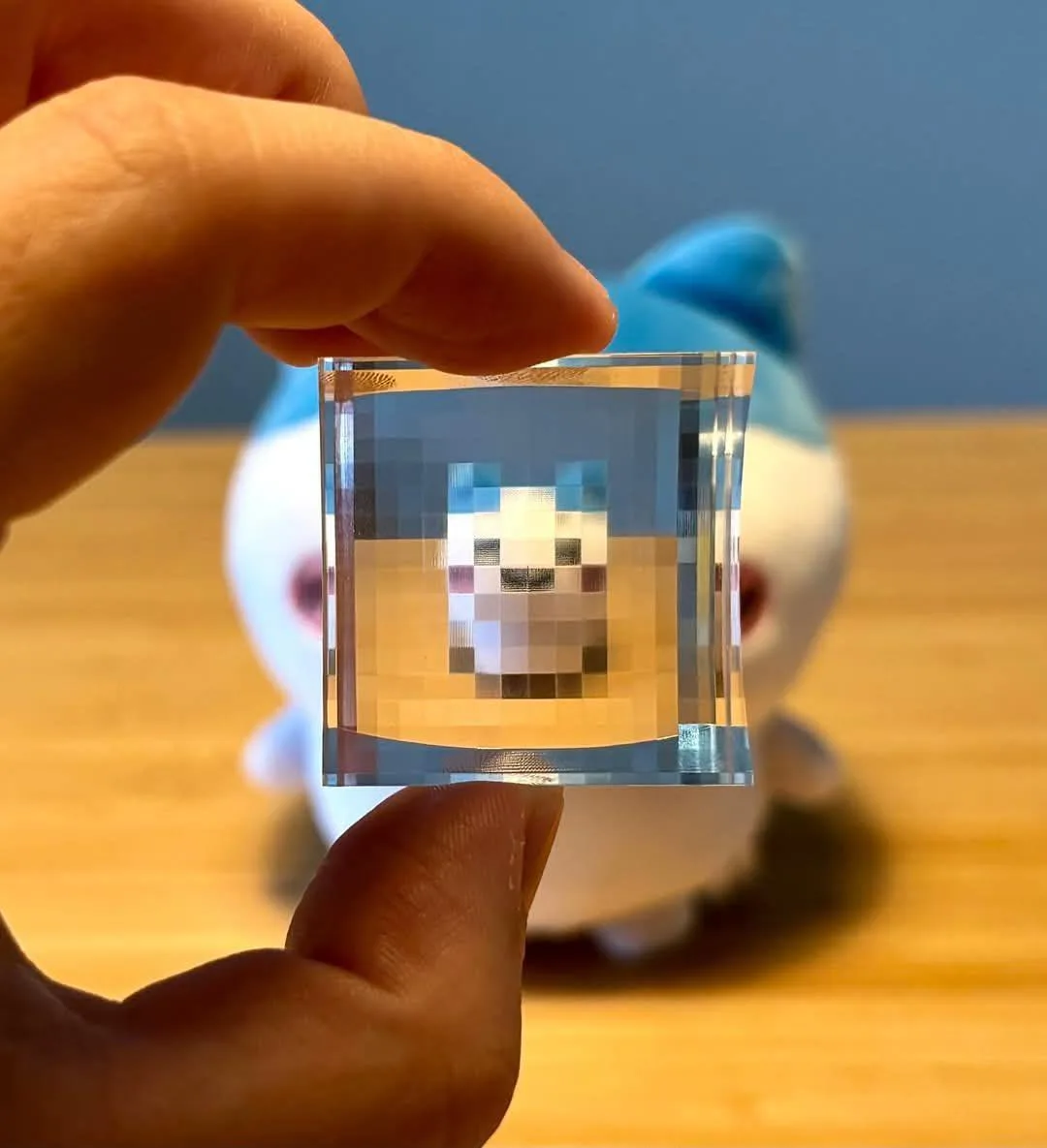 A hand holds a small, clear glass cube that refracts light to reveal a pixelated, blocky face inside. Behind the cube, a…
