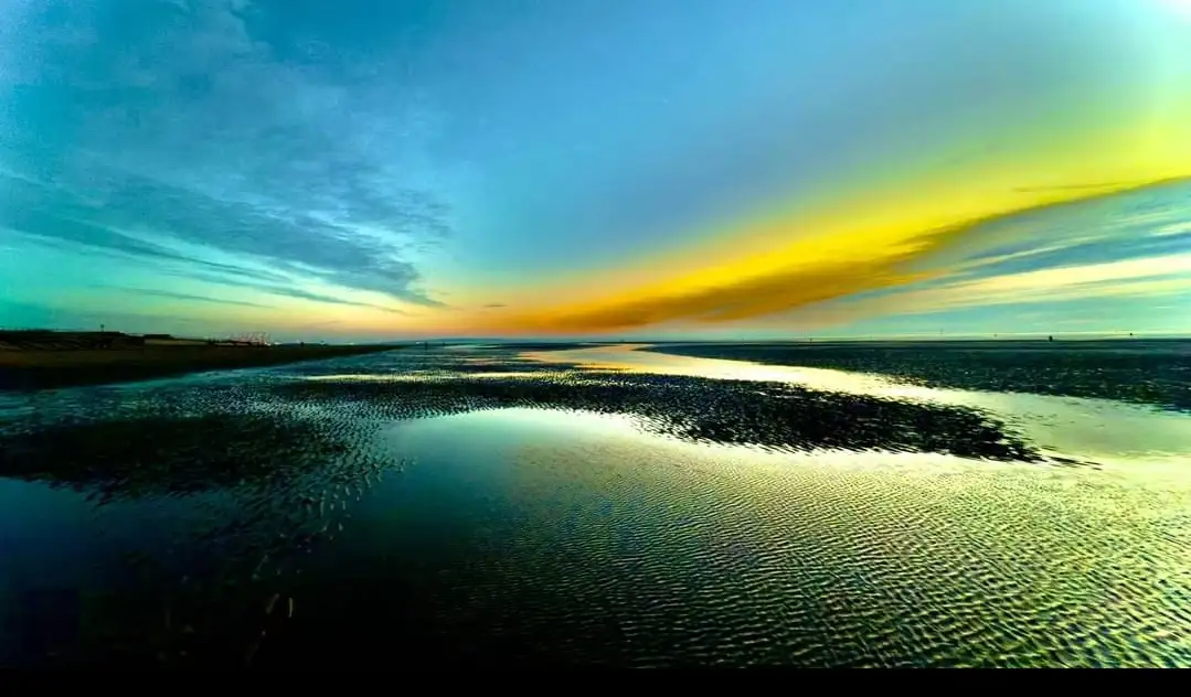 A serene landscape photo of a tidal flat at sunset, with rippling water reflecting vibrant yellow and blue skies. The…