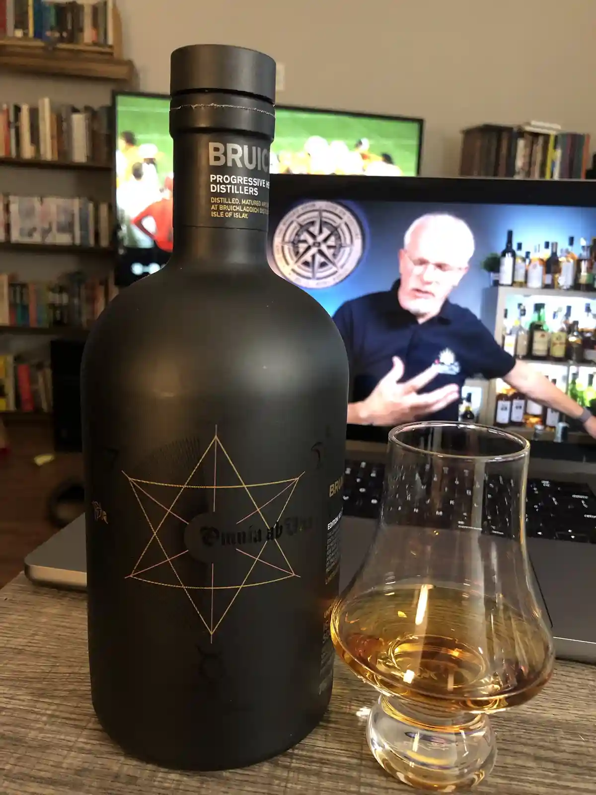 A black bottle of Bruichladdich 'Omnia ab Uno' whisky sits next to a tasting glass filled with amber liquid on a wooden…