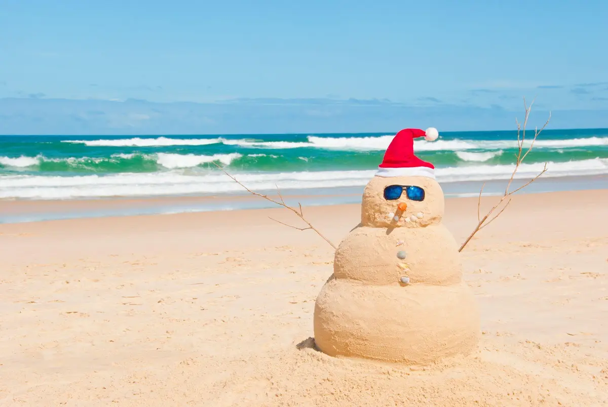 A sand sculpture shaped like a snowman stands on a sunny beach, wearing a red Santa hat, blue sunglasses, and using…