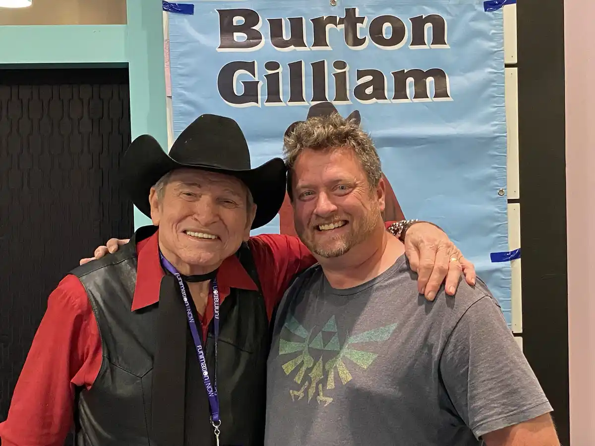 Two men are posing for a photo in front of a blue banner that reads 'Burton Gilliam'. The older man on the left wears a…