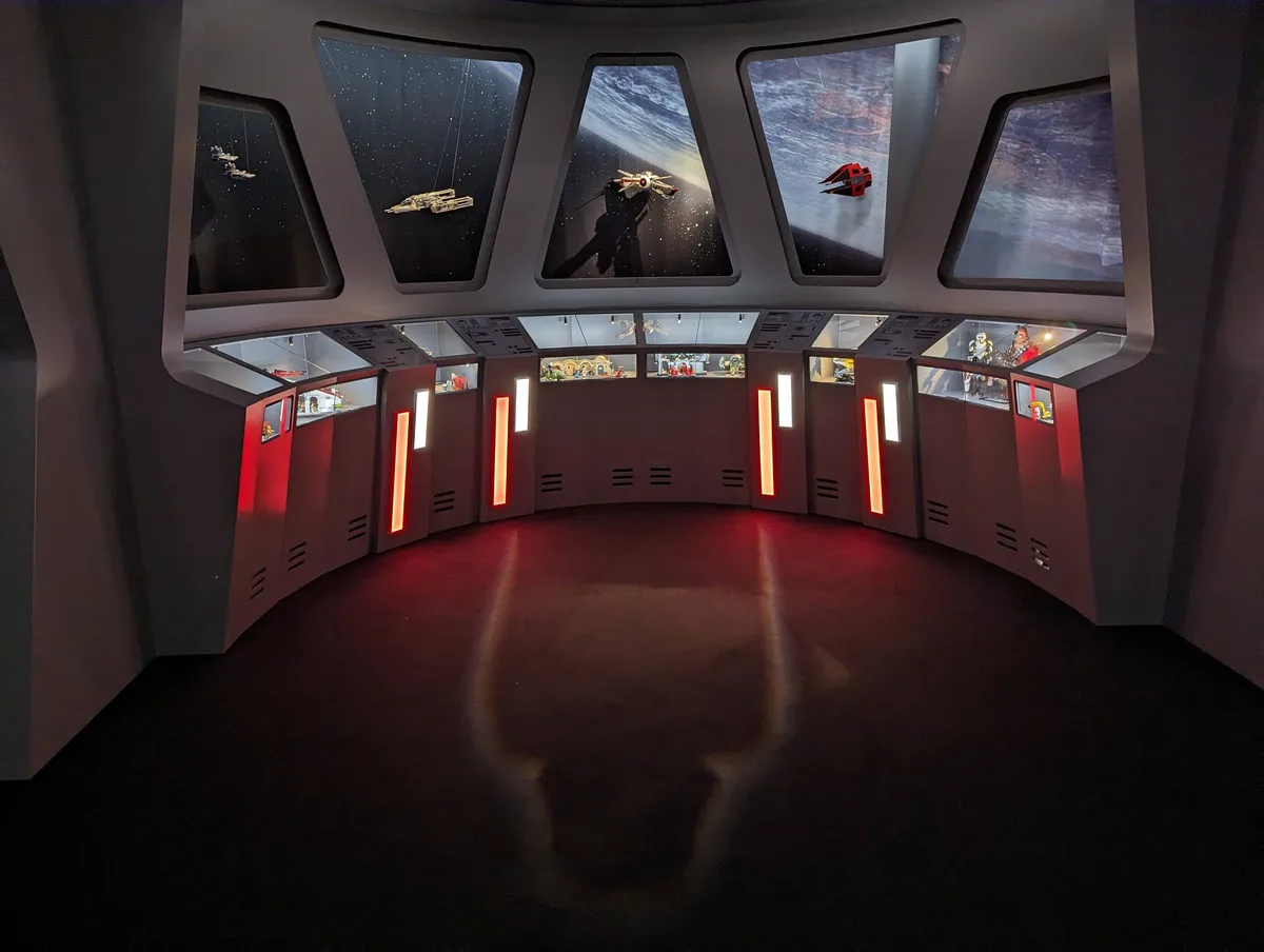 A photo of an empty, curved Star Wars-themed exhibit room with large windows showing space scenes and model ships, and…