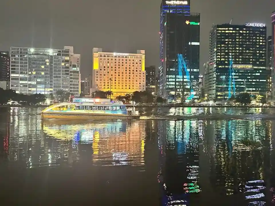 A brightly lit yellow and blue water taxi boat moves across a river at night, with its lights reflecting on the water…