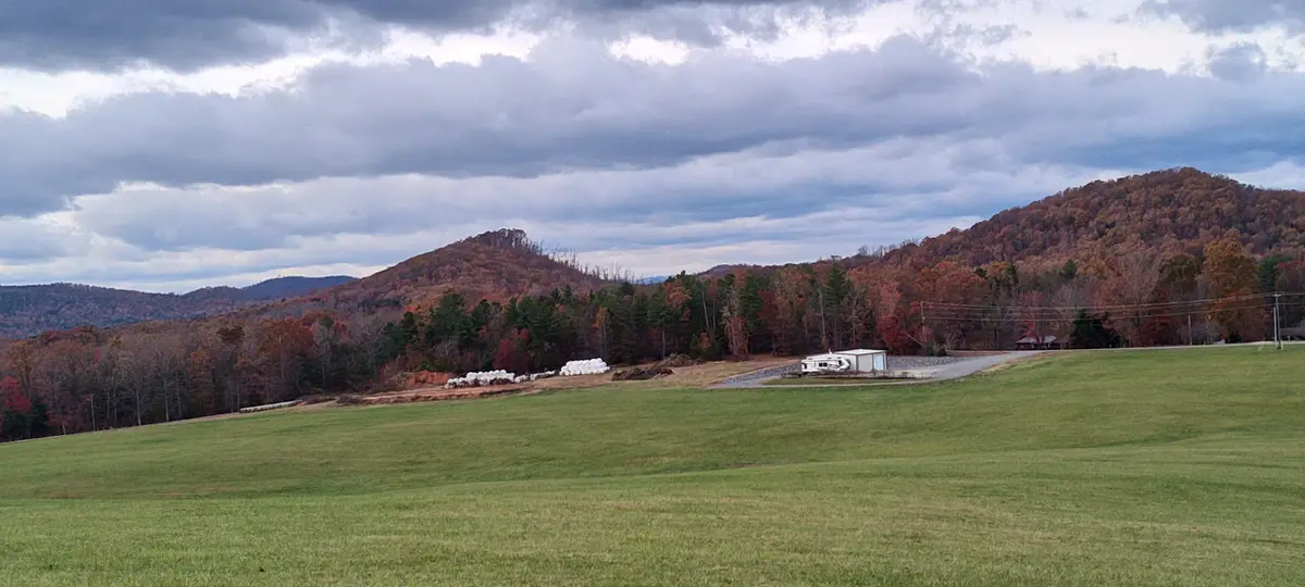 A wide landscape photo shows a green field in front of autumn-colored hills under a cloudy sky, with a white RV parked near…