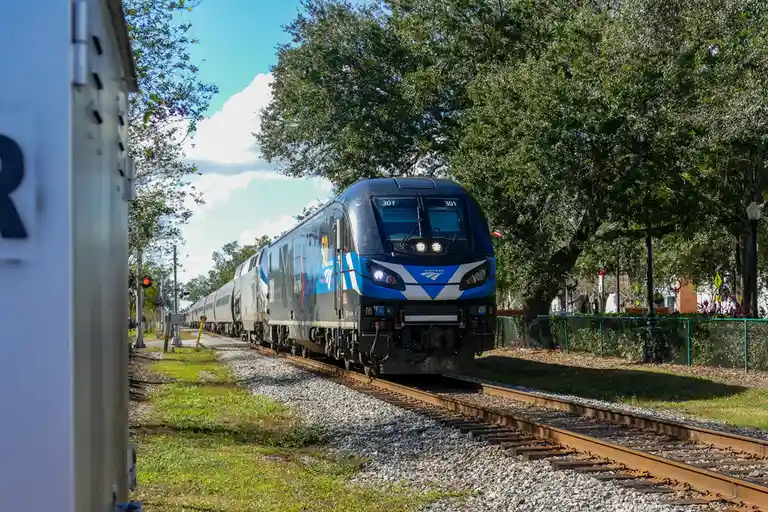 An Amtrak train, number 301, is shown moving along tracks under a partly cloudy sky, flanked by trees and a fence. The…