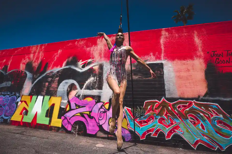 A person in a sparkly, fringed costume and ballet shoes is suspended mid-air by a harness attached to a pole, striking a…