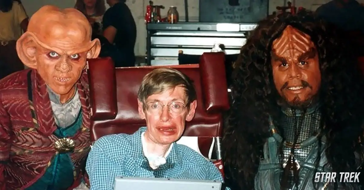 A photo shows physicist Stephen Hawking seated in a red chair, flanked by two Star Trek alien actors in full costume. The…