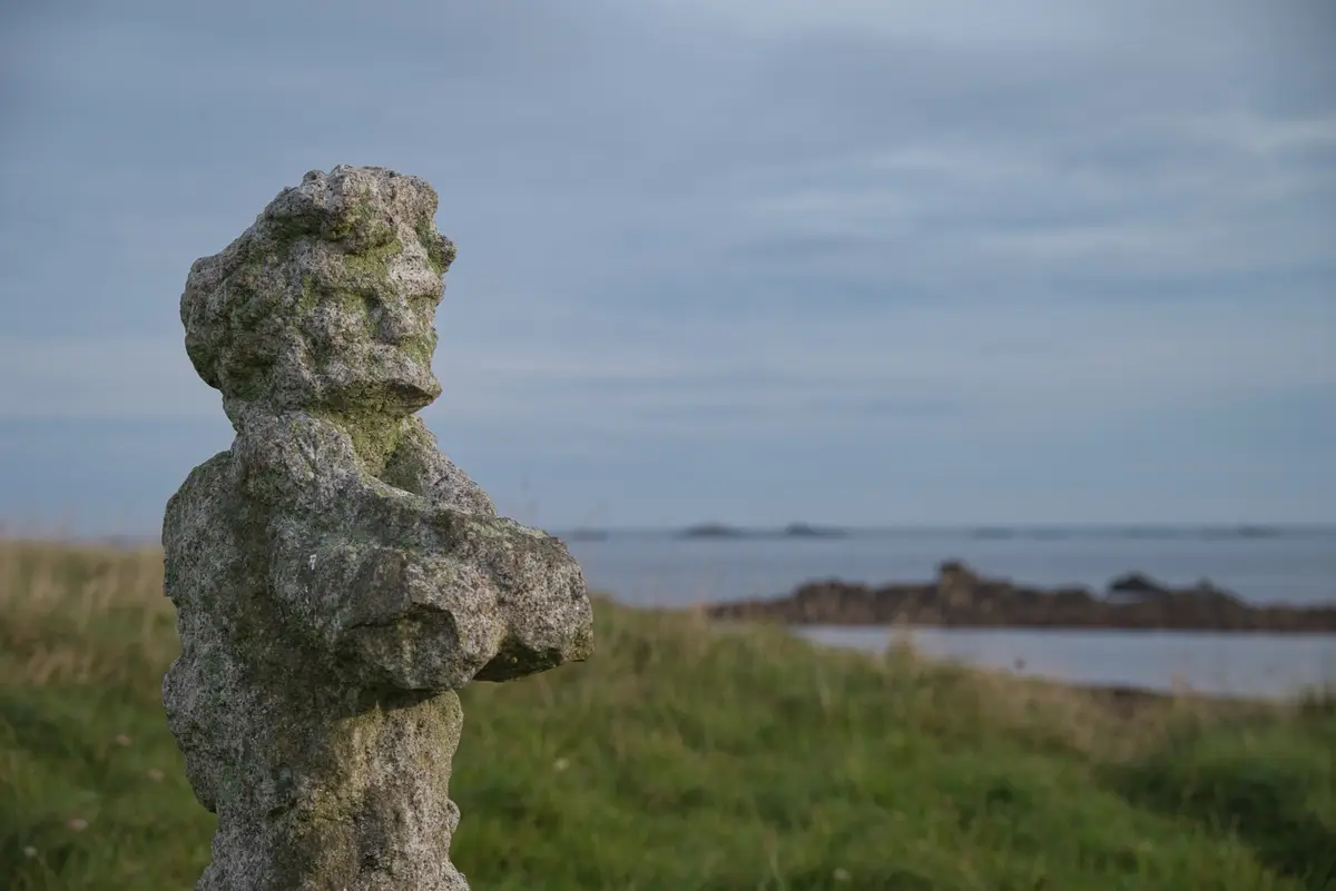 A weathered, moss-covered stone statue of a person stands in a grassy field near the sea, looking out toward the horizon…