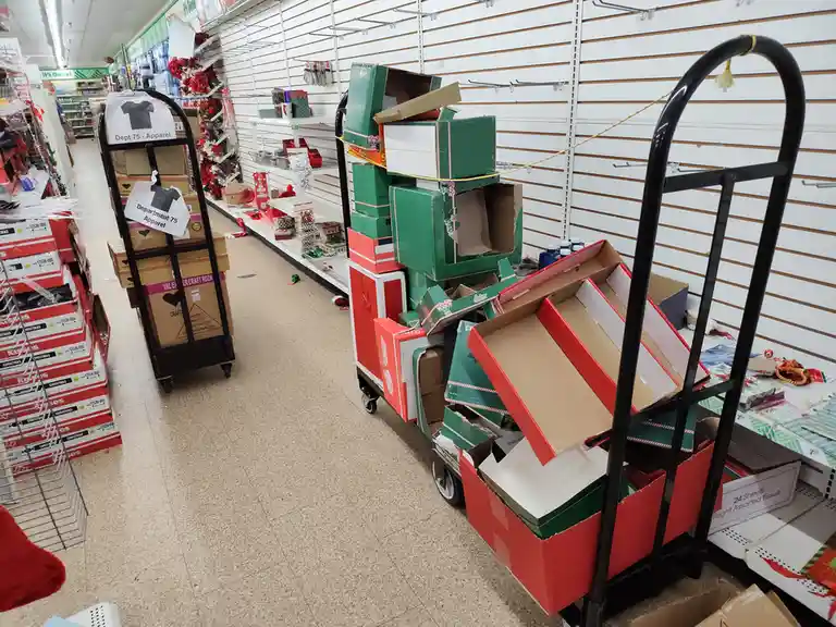 A store aisle with empty shelves and carts piled high with red and green cardboard boxes, some labeled 'Department 75…