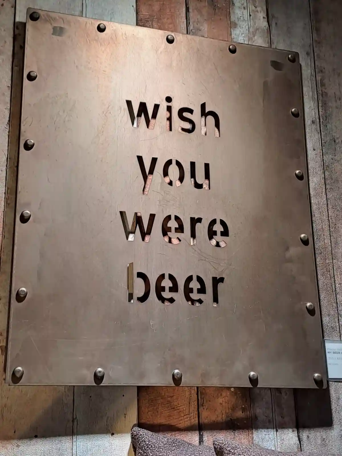 A close-up of a rustic metal plaque mounted on a wooden wall, featuring the phrase 'wish you were beer' cut out in bold…