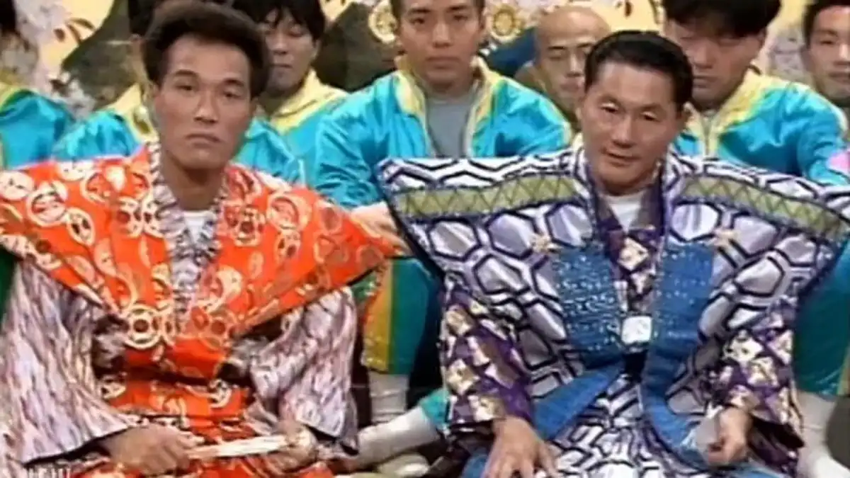 Two men in brightly patterned, oversized, and mismatched traditional-style garments sit side by side, looking unimpressed…