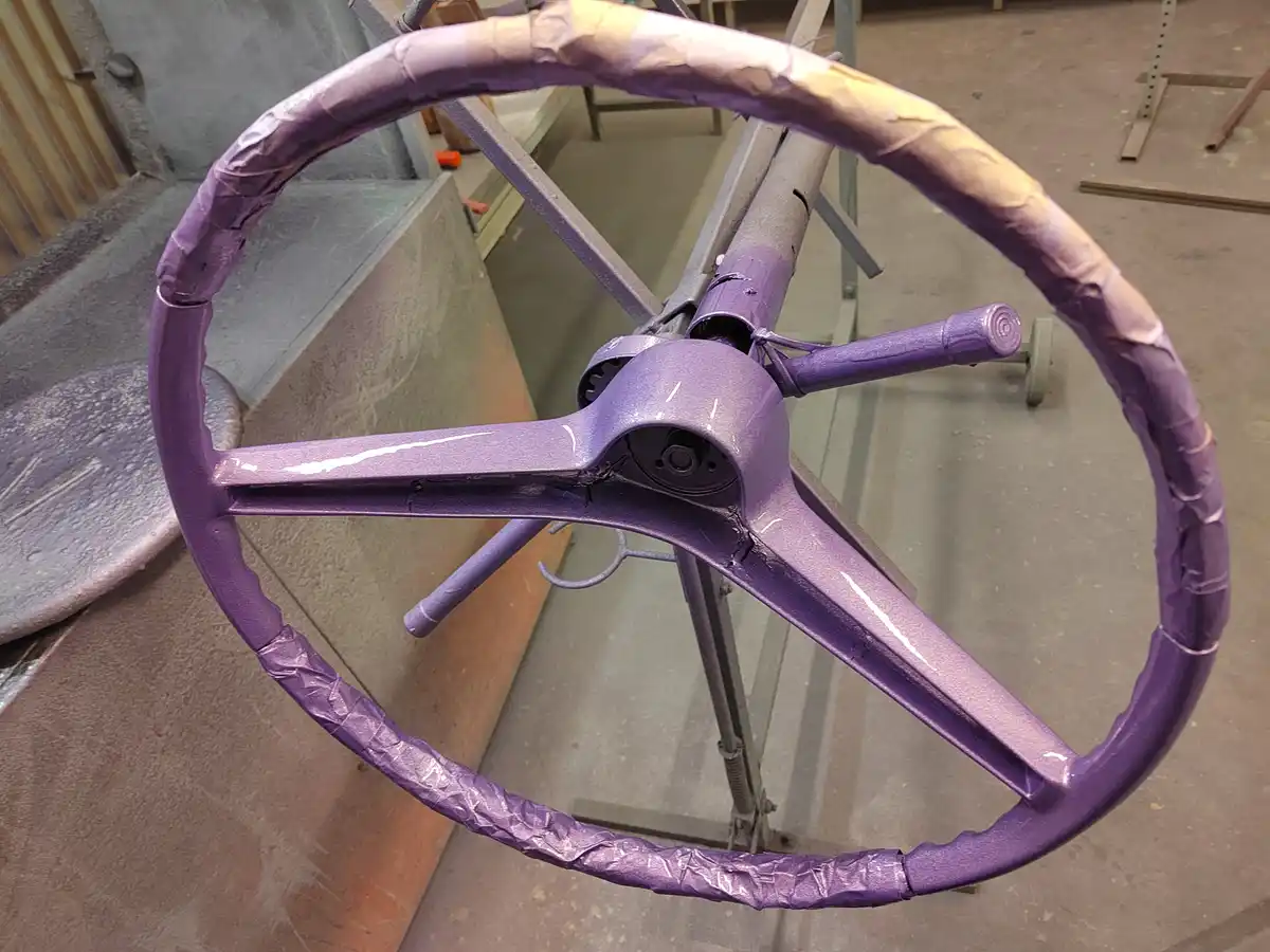 A close-up photo shows a large, metallic steering wheel freshly painted a vibrant purple, with protective tape still wrapped…