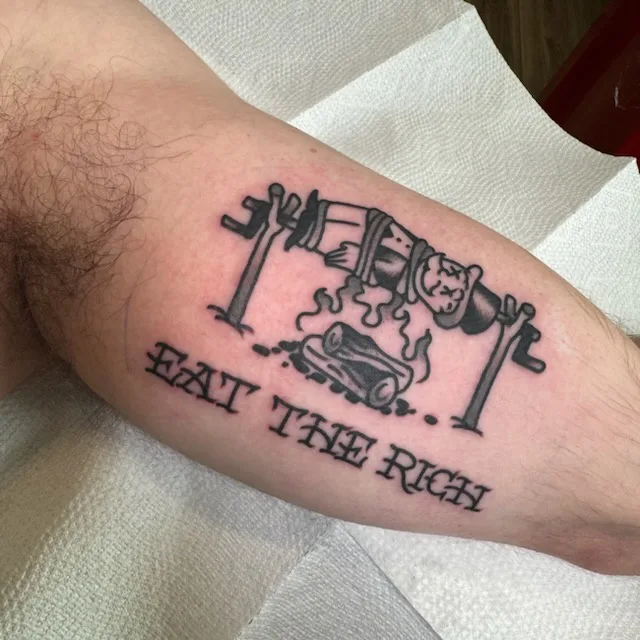 A black-and-white tattoo on a person's forearm depicts a cartoonish cannon firing a log, with the text 'EAT THE RICH'…