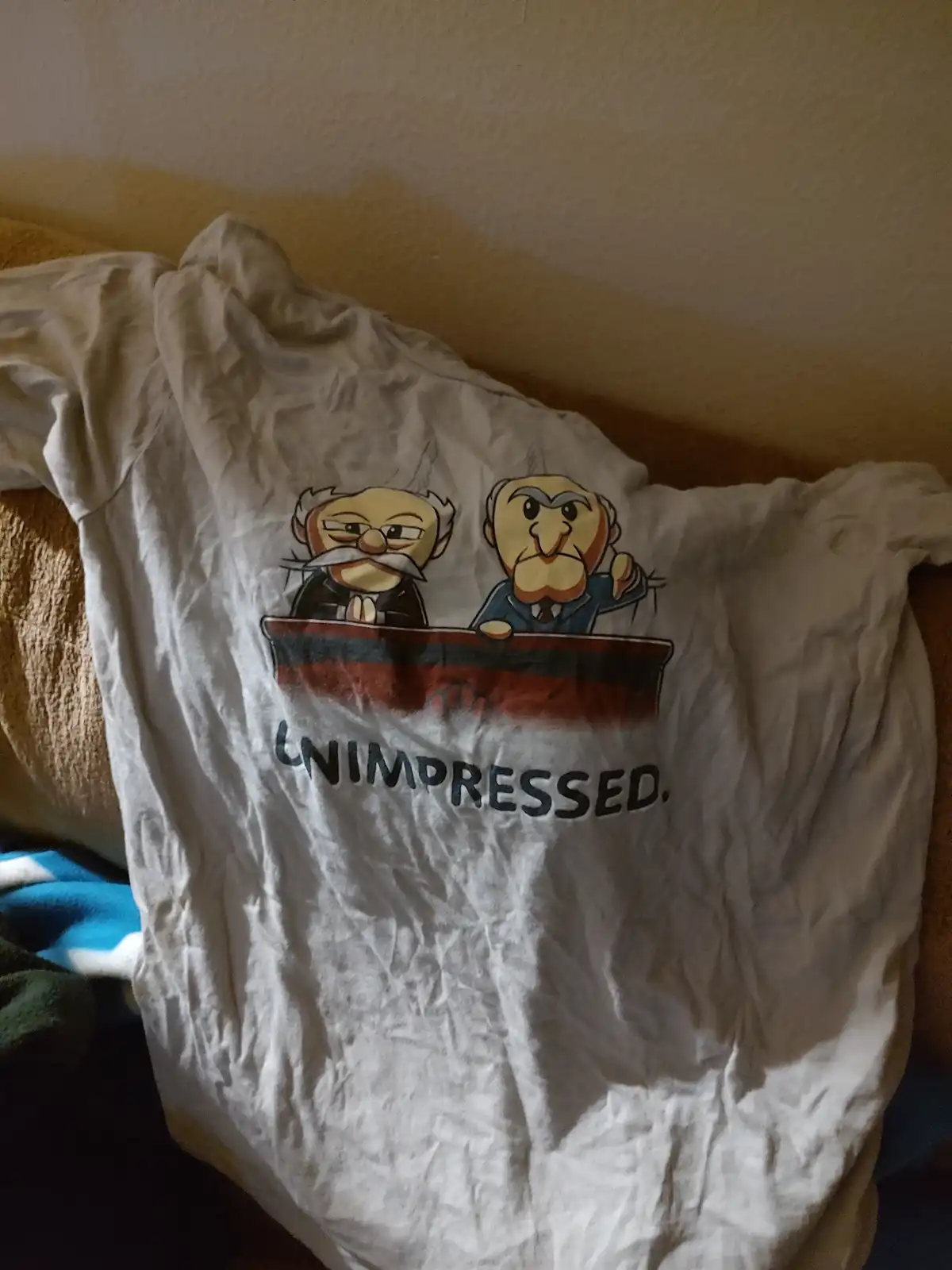 A wrinkled white t-shirt displays a cartoon graphic of two stern-looking, bearded men sitting behind a desk, with the word…