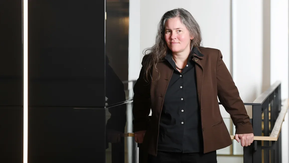 a woman with long gray hair wearing a brown blazer over a black shirt stands leaning on a railing in a modern building…