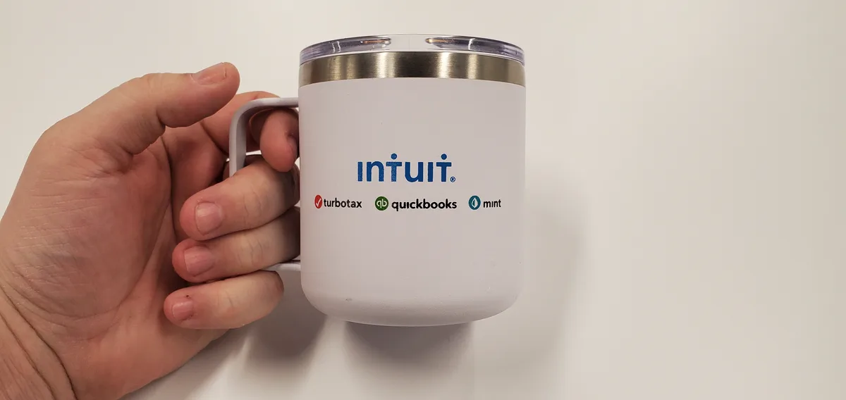 A hand holds a white insulated mug with a clear lid and silver rim. The mug displays the word 'intuit.' in blue, followed by…