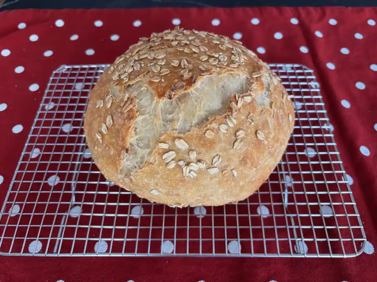A round loaf of bread with a cracked, uneven surface resembling a face, sits on a metal cooling rack atop a red cloth with…