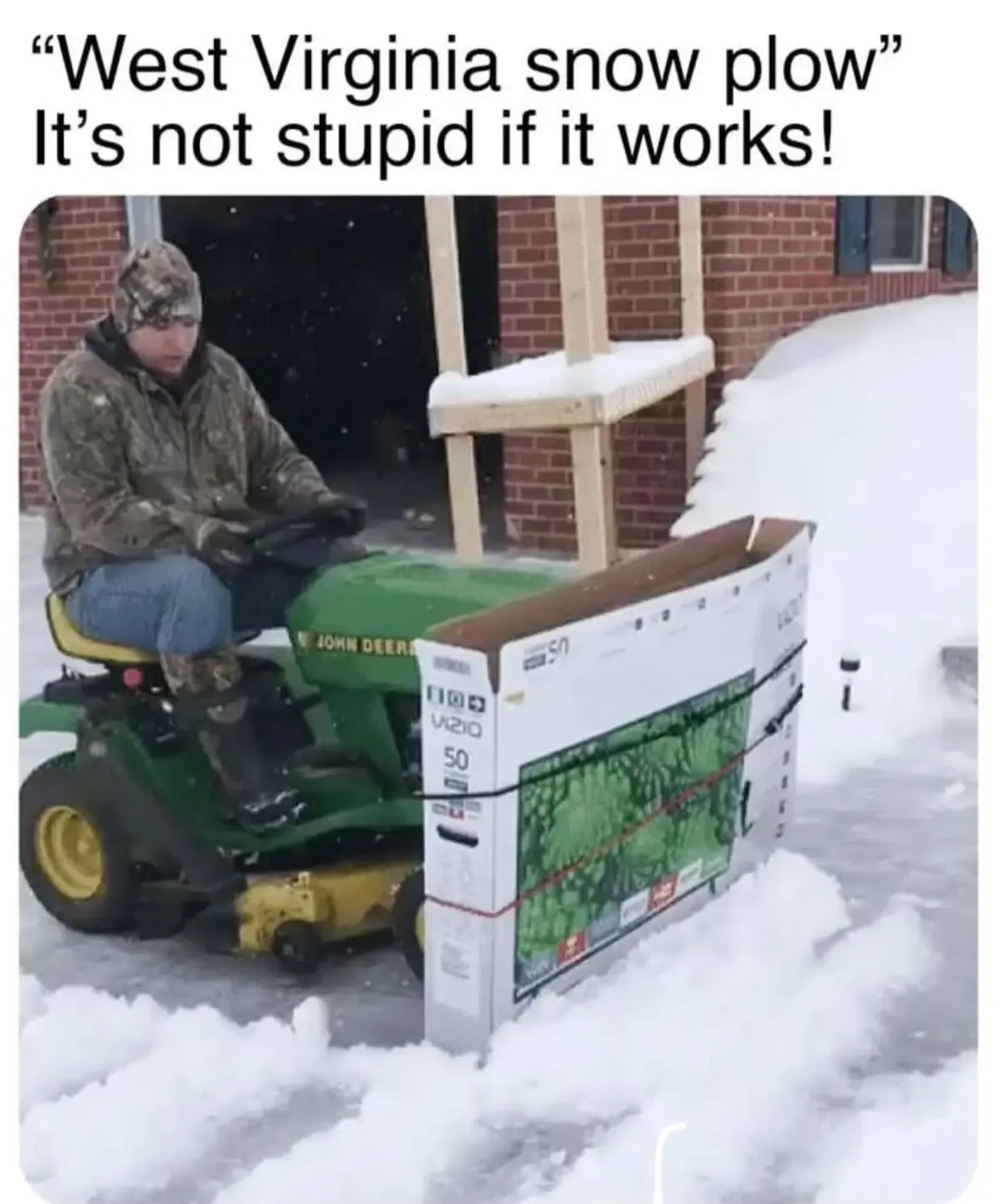 A person in camouflage gear sits on a green John Deere riding lawnmower in a snowy yard, pushing a large, flat-packed TV box…