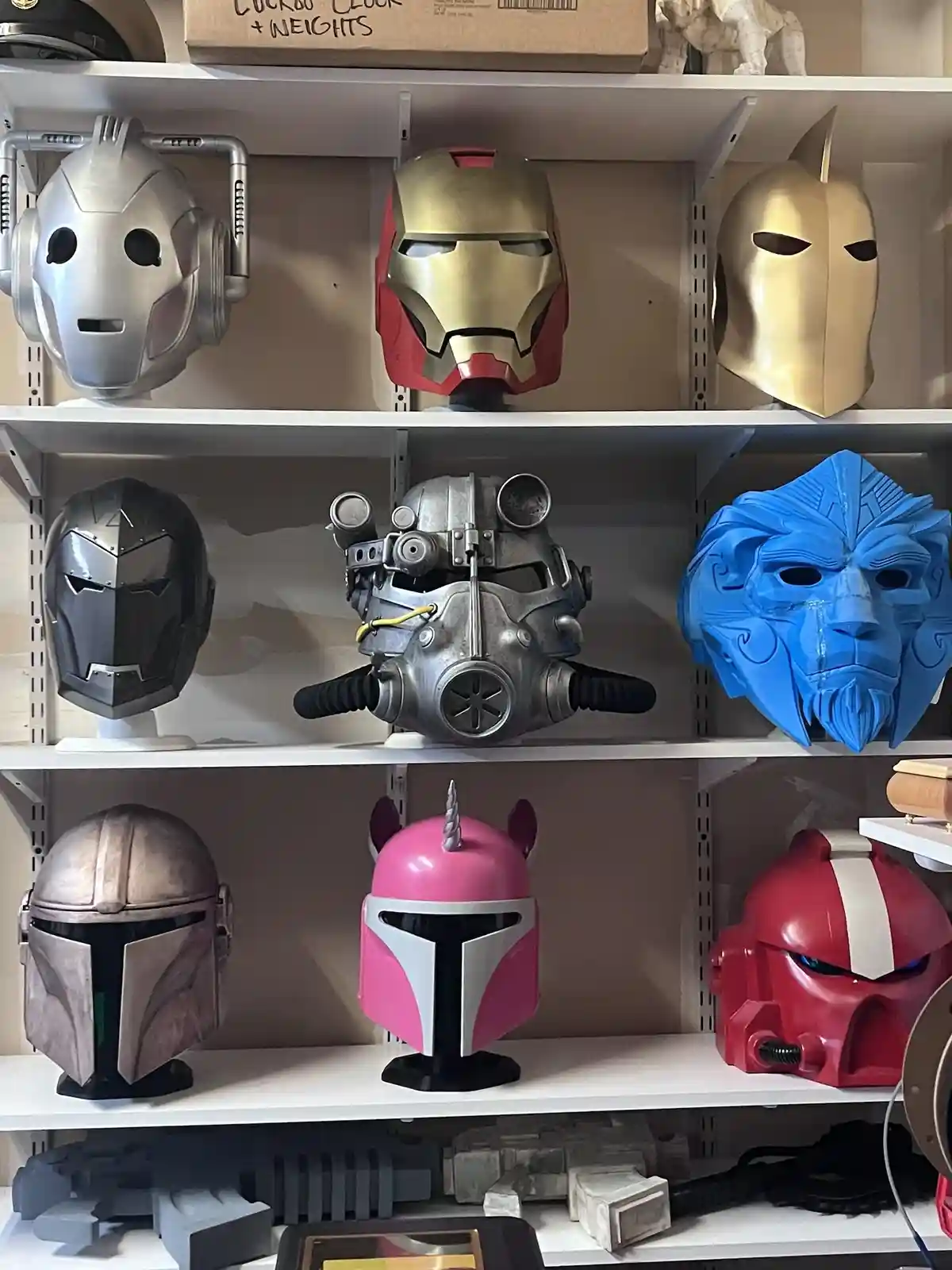 A white shelving unit displays nine different custom or replica helmets and masks from various fictional universes…