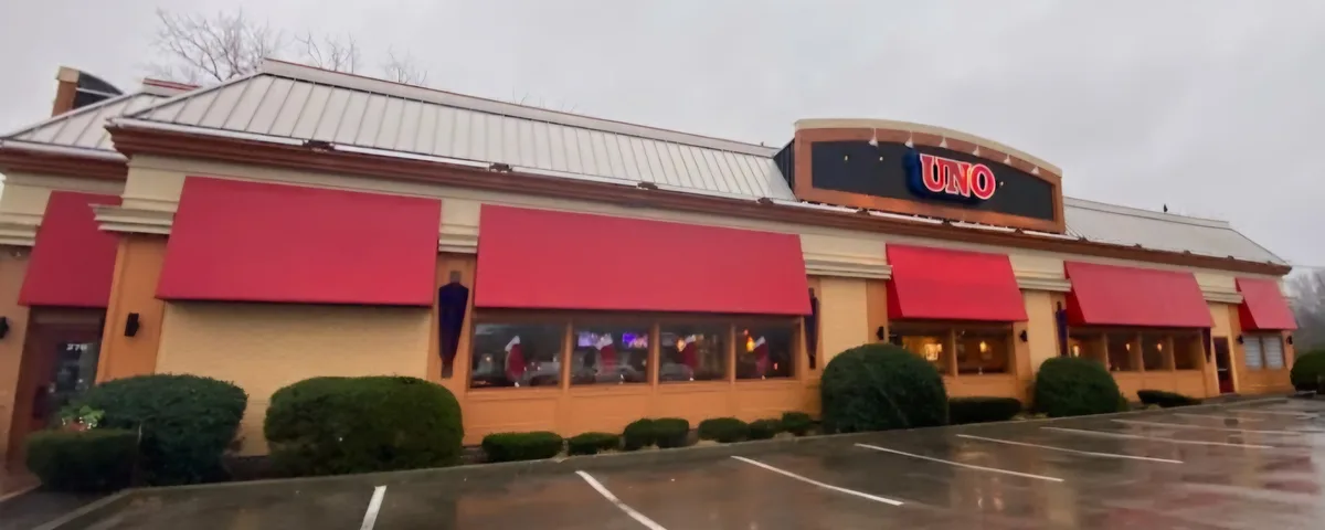 An exterior shot of a restaurant with a beige brick facade, red awnings, and a large illuminated sign that reads 'UNO' above…