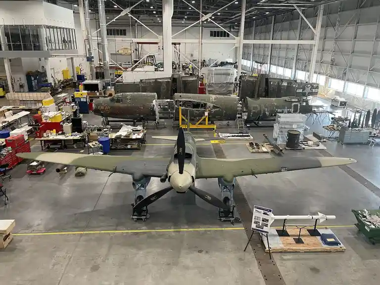 A wide-angle photo inside a large aircraft hangar shows several military planes in various stages of restoration or…