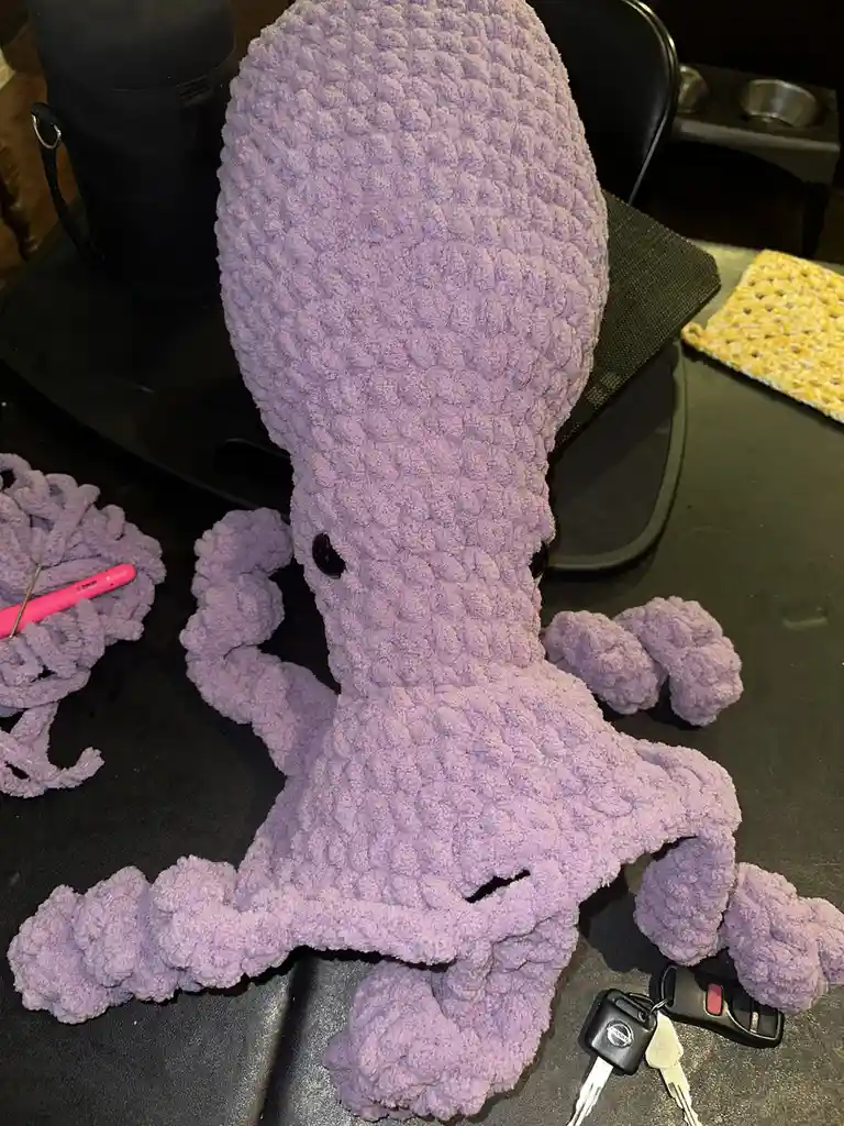 A large, purple, crocheted octopus plushie sits on a black office chair, with a pink crochet hook and yarn pile nearby. A…