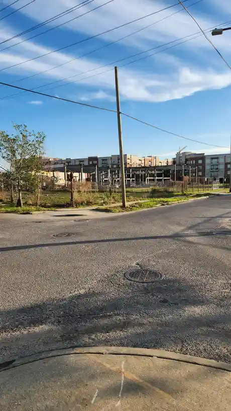 A photo of an asphalt street with manholes and a utility pole, leading to a fenced construction site with partially built…