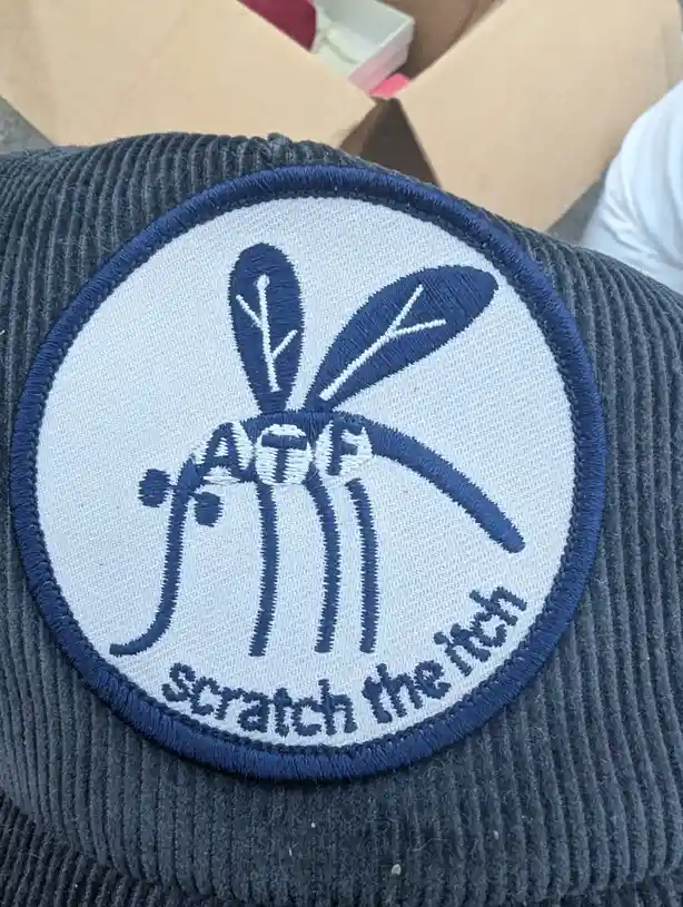 A close-up of a circular embroidered patch on a dark gray corduroy trucker hat, featuring a stylized blue mosquito with…