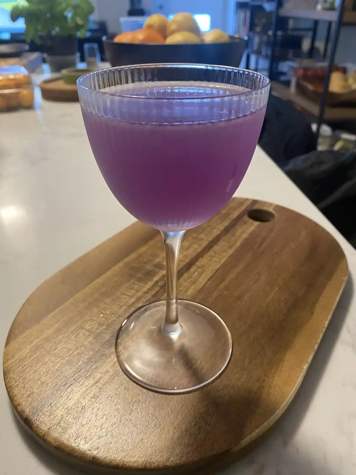 A close-up photo shows a purple cocktail in a stemmed glass with a ribbed rim, resting on a wooden board atop a kitchen…