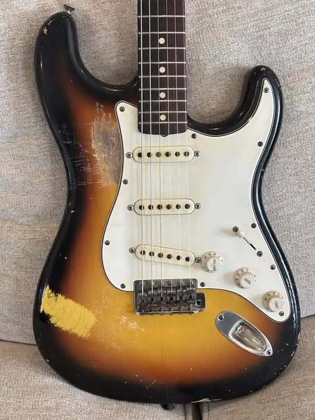 A heavily worn, sunburst-finish electric guitar with visible scratches, dents, and faded paint lies on a textured fabric…