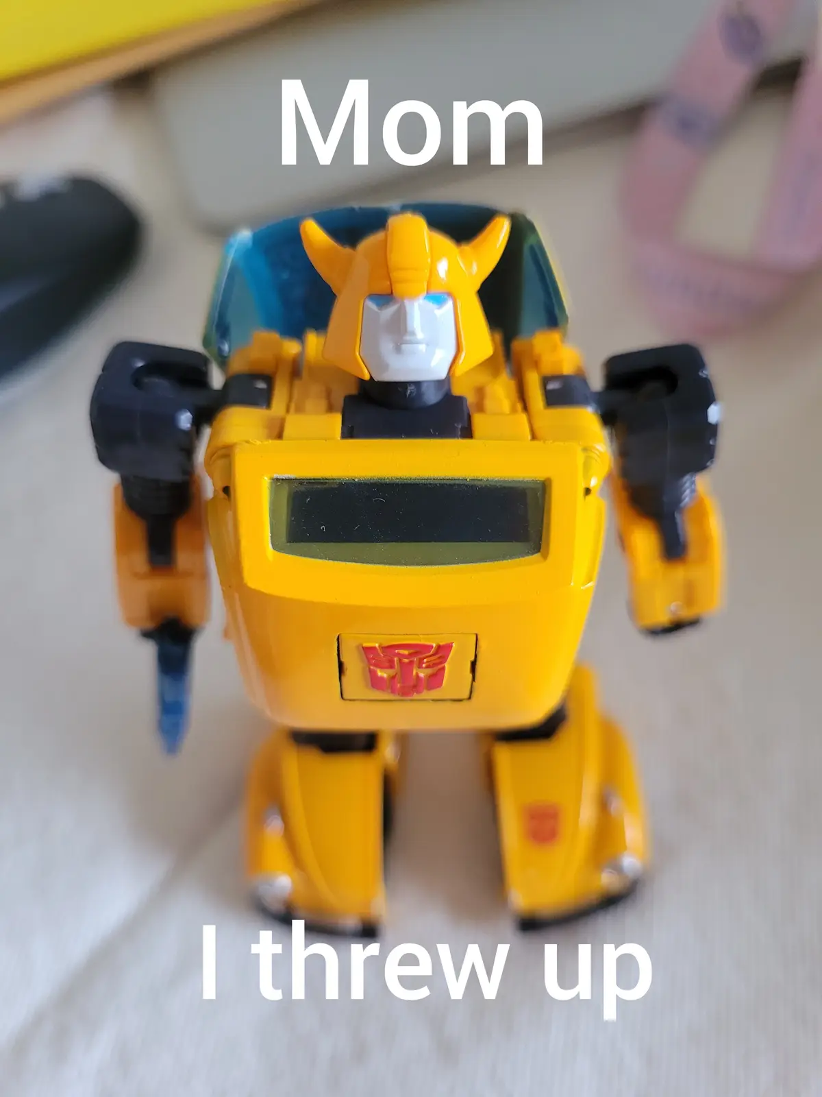 A yellow Transformers toy robot, resembling Bumblebee, stands on a light-colored surface with the word 'Mom' overlaid at the…