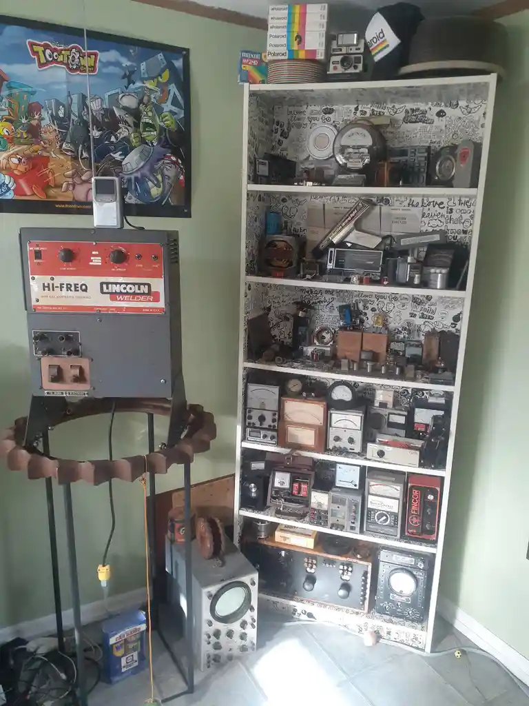 A room filled with vintage electronics, including a Lincoln Hi-Freq welder on a stand, a tall white shelf packed with analog…