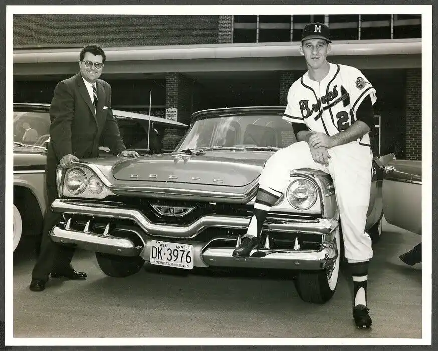 A black-and-white photo shows a man in a suit and glasses leaning on a vintage Dodge car while a baseball player in a Braves…