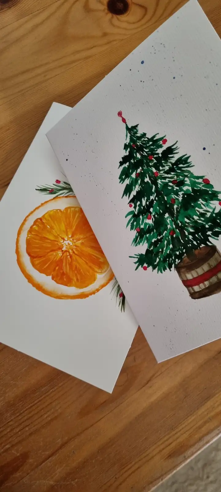 Two watercolor holiday cards lie on a wooden surface; one depicts a sliced orange with greenery, the other shows a green…