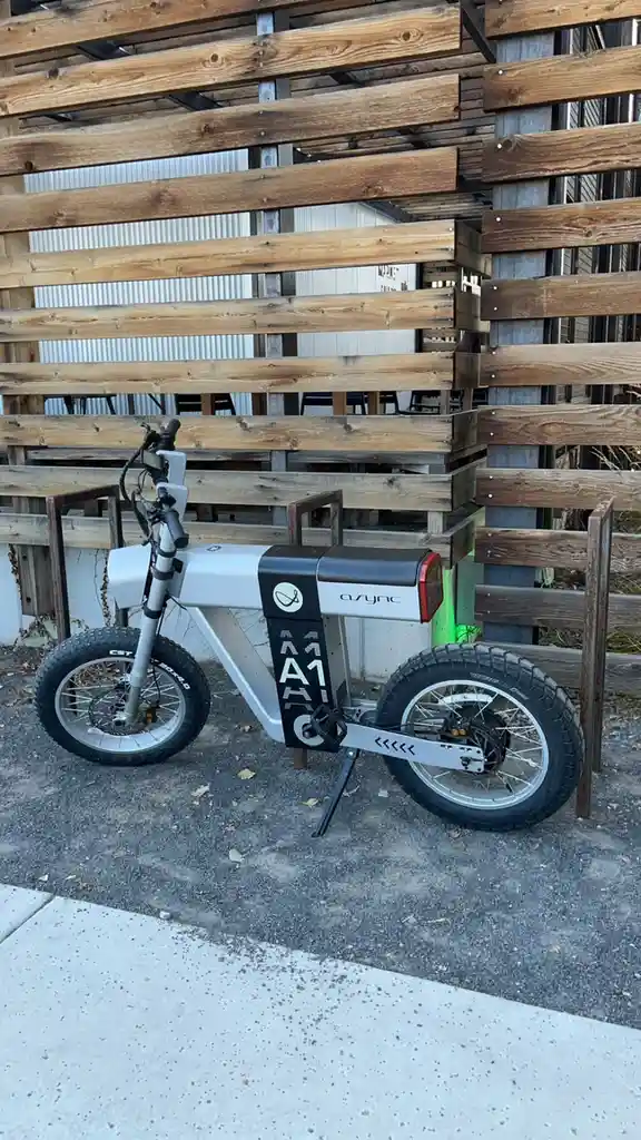 A silver and black electric bicycle branded 'async' is parked on gravel next to a wooden slat fence. The bike's black frame…