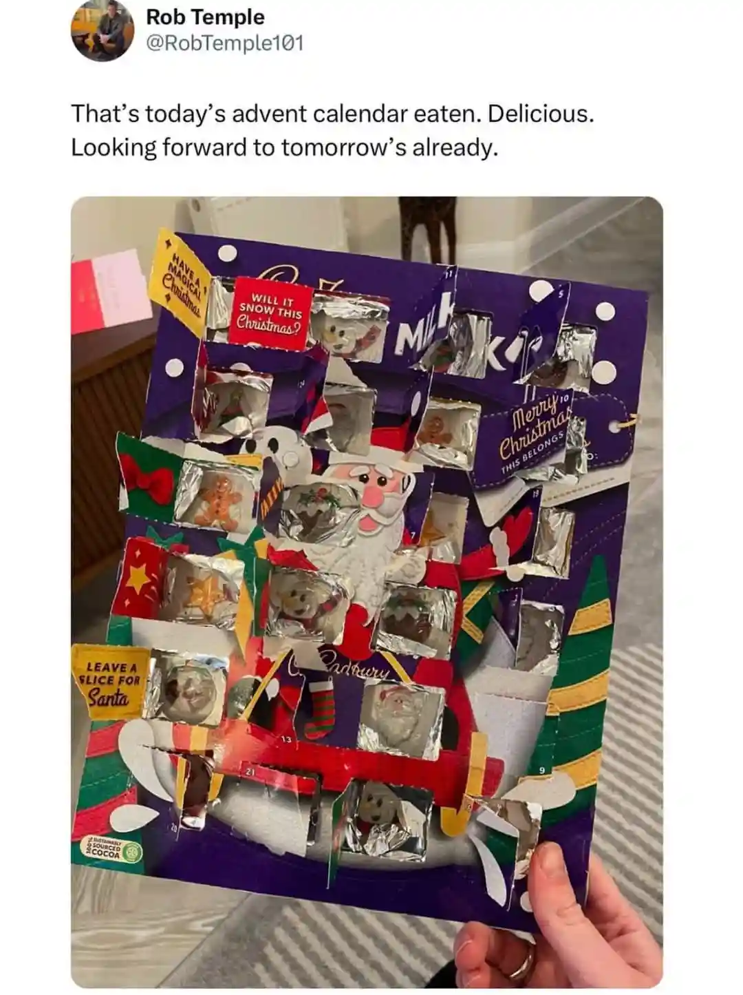 A hand holds a Cadbury advent calendar with most of its doors torn open, revealing foil-wrapped chocolates inside. The…