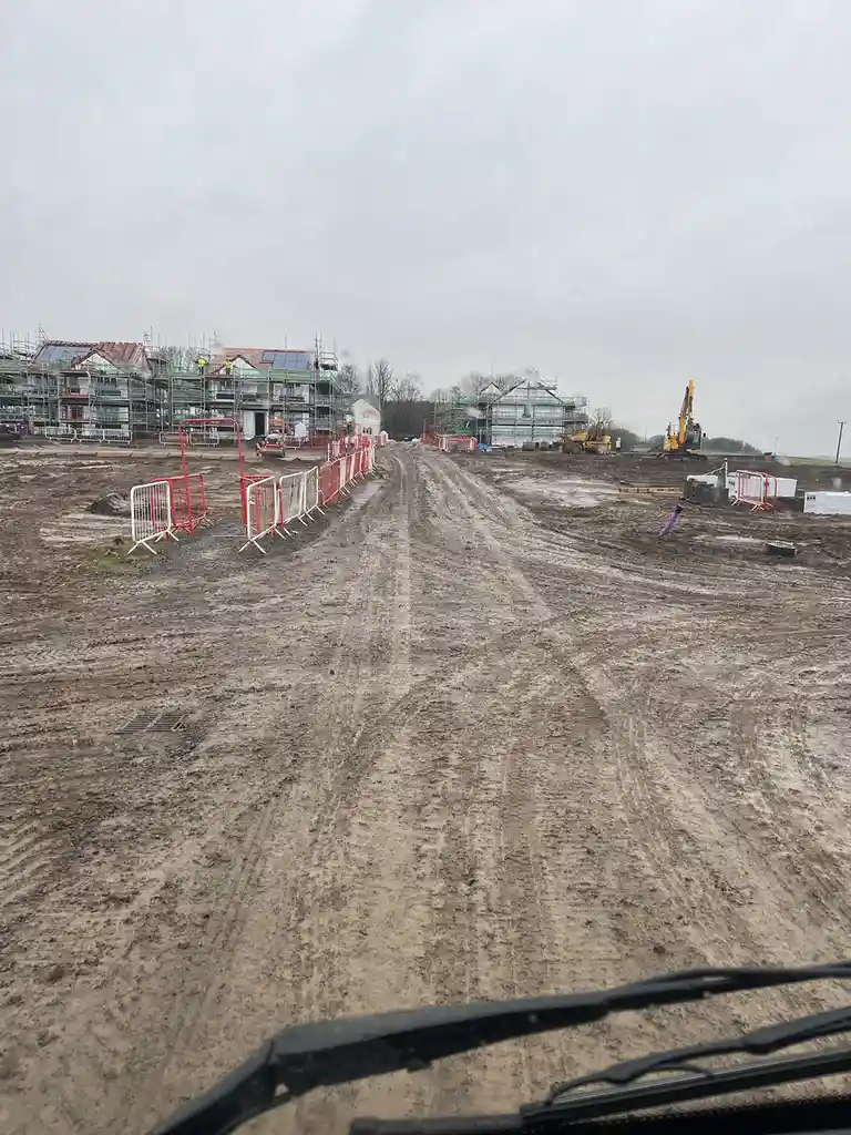 A muddy construction site with partially built houses covered in scaffolding, viewed from inside a vehicle with wipers…