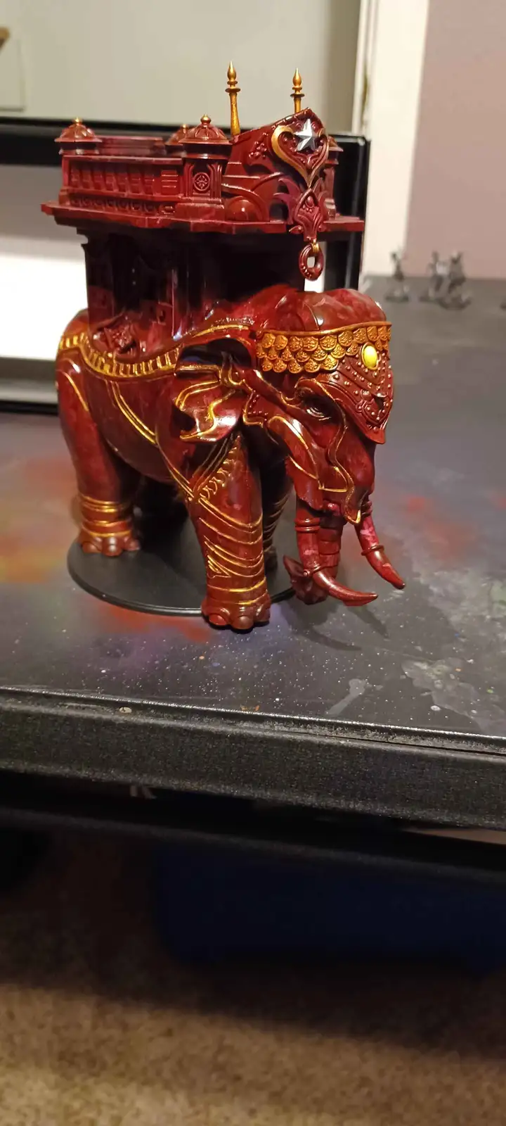 A highly detailed, hand-painted miniature of a red elephant adorned with gold trim and an ornate, palace-like howdah on its…